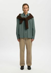 Jeune homme portant une chemise verte boutonnée, un pantalon beige, des chaussures foncées et un pull marron posé sur les épaules, se tenant devant un fond blanc.