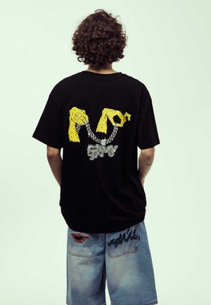 Camiseta negra con un gráfico amarillo de manos y una cadena plateada, con la palabra "Grimy" en una fuente decorativa en la parte posterior.