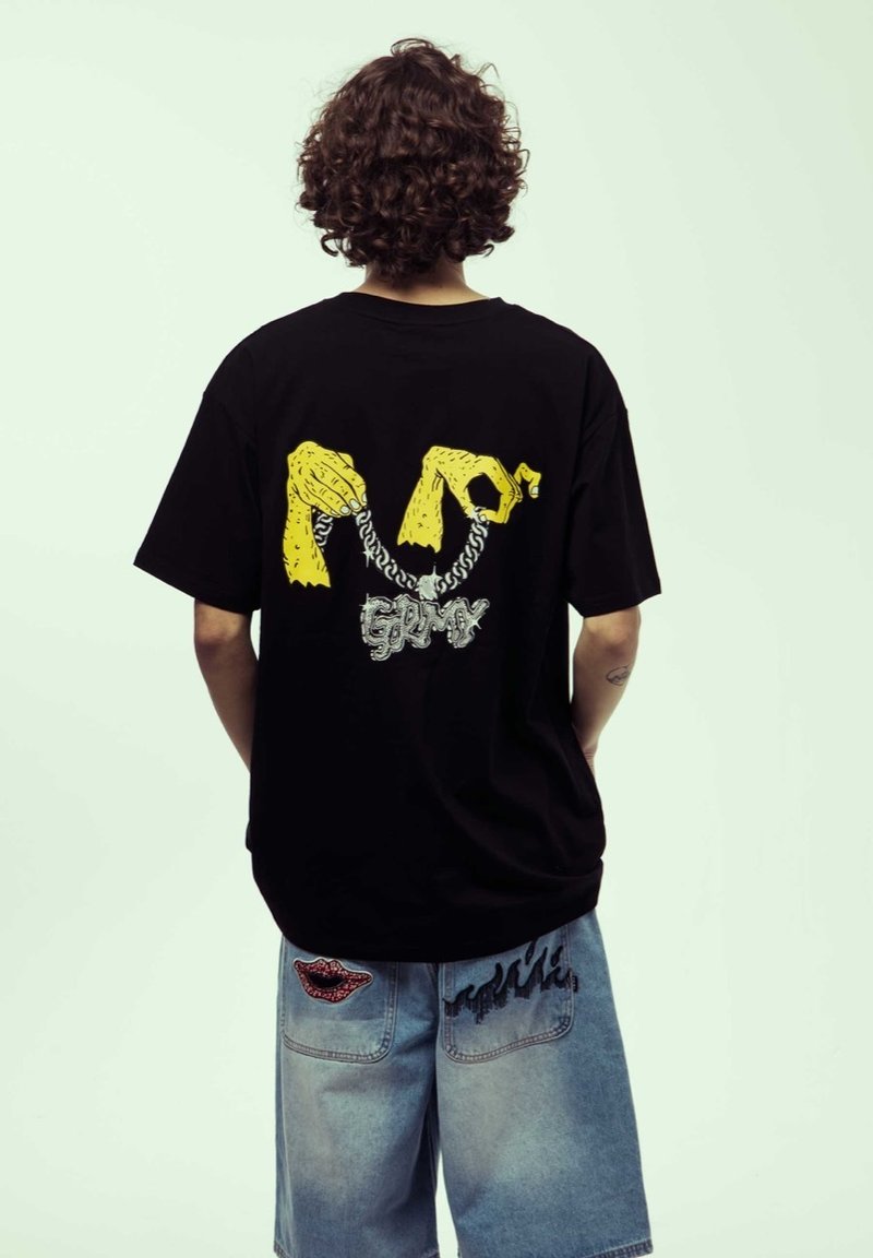 T-shirt nera con una grafica gialla di mani e una catena d'argento, con la parola "Grimy" in un font decorativo sul retro.