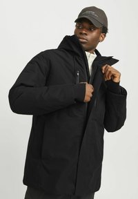 Schwarze isolierte Jacke mit hohem Kragen und Kapuze, ausgestattet mit einem frontalen Reißverschluss und zwei Reißverschlusstaschen auf der Brust. Glatte, matte Stoffoberfläche.