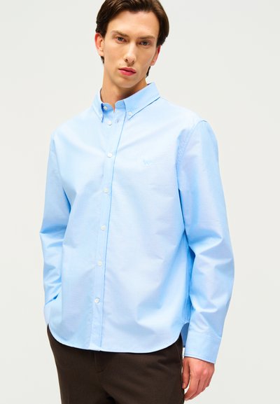 Νεαρός άνδρας με ανοιχτό μπλε πουκάμισο με μακριά μανίκια και σκούρο παντελόνι, στέκεται μπροστά σε μια απλή ανοιχτόχρωμη επιφάνεια.