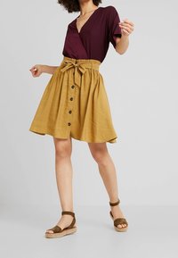 Burgundy wrap top with V-kaula, yhdistettynä sinapinkeltaiseen, polvipituiseen hameeseen, jossa on nauhavyö ja tummat napit edessä.