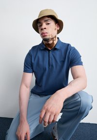 Námořnická modrá polo tričko s límečkem a třemi knoflíky, spárované se světle modrými kalhotami a béžovým kloboukem typu bucket.