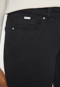 Jeans de mezclilla negra con un ajuste slim, que cuentan con herrajes plateados, bolsillos frontales y una pequeña etiqueta con el logotipo en el bolsillo derecho. Textura suave.