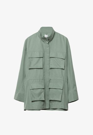 Frau trägt große, braun getönte Sonnenbrillen und eine hellgrüne Jacke mit mehreren Klappentaschen und einem Taillenbindegürtel.