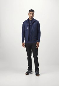 Tamsiai mėlynas lengvas švarkelis su užtrauktuku priekyje ir gobtuvu, dėvimas ant tamsiai mėlynos polo marškinėlių. Juodos kelnės ir daugių spalvų sportbačiai užbaigia aprangą.