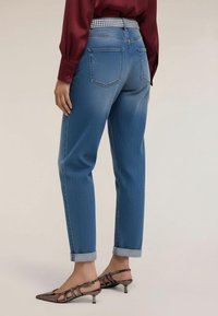 Osoba ubrana w niebieskie dżinsy z mankietami, bordową koszulę z długim rękawem i buty na niskim obcasie z zaokrąglonym noskiem, widziana od tyłu.