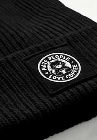 Black knit beanie featuring a textured design. A round patch displays a cartoon cat with the text "HATE PEOPLE, LOVE COFFEE."