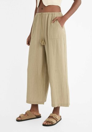 Pantalons larges beige texturés avec une taille élastique, cordon de serrage avec des pompons et poches latérales. Associés à des sandales à boucle couleur fauve.