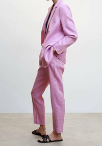 Różowy lniany garnitur składający się z dopasowanej marynarki z guzikami i pasujących spodni. Noszony z czarnymi sandałami na cienkich paskach. Minimalistyczny design.