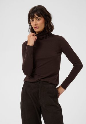 Femme aux cheveux ondulés foncés portant un col roulé marron foncé et un pantalon, debout avec une main sur la hanche et l'autre touchant le col.
