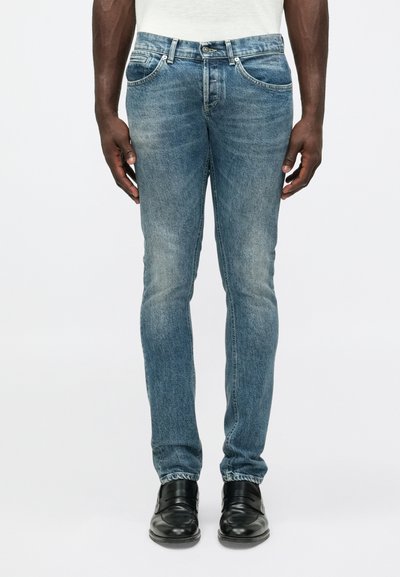 Blaue Denim-Jeans mit schlanker Passform, mit verwaschenem Finish, Fünf-Taschen-Design und metallischen Beschlägen an der Taille. Getragen mit schwarzen Schuhen.