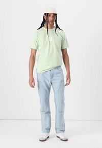 Lichtgroene katoen T-shirt met witte print, gecombineerd met lichtblauwe denim jeans met zijnaden en voorzakken, gedragen met een beige pet en sneakers.
