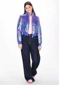 Irydescencyjna krótka kurtka puchowa w kolorze fioletowym z wysokim kołnierzem, zapinana na zamek, z dwoma przednimi kieszeniami, w połączeniu z ciemnymi spodniami typu wide-leg.