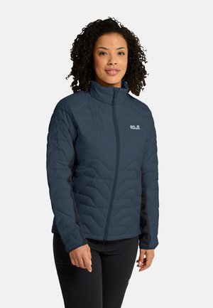 Frau mit lockigem Haar, die eine dunkelblaue gesteppte Jacke mit Reißverschluss und schwarze Hosen trägt, steht vor einem einfarbigen Hintergrund.
