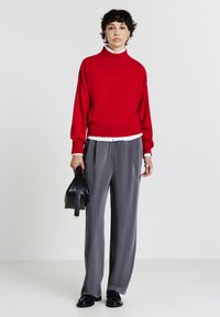 Maglione rosso a maglia con collo alto, abbinato a pantaloni grigi a gamba larga. Il modello tiene una piccola borsa nera e indossa scarpe nere.