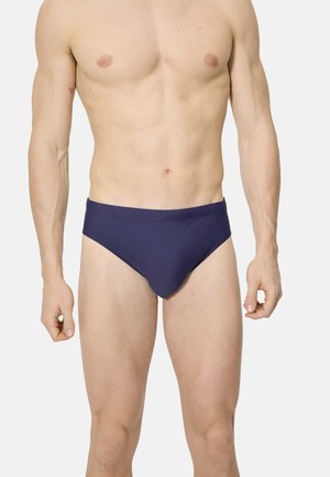 Hombre con bañador corto azul oscuro liso, de pie con los brazos relajados contra un fondo claro y liso.