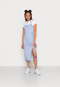 Vestido floral azul com textura franzida, fenda lateral e laço ajustável. Usado sobre uma t-shirt branca, combinado com ténis leves.