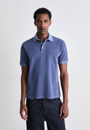 Polo shirt in light blue cotton with a classic collar, two-button placket, and a small embroidered logo on the left chest, paired with dark jeans.