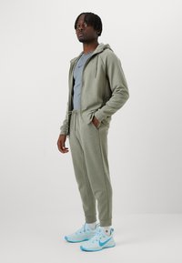 Light grey tracksuit with a zip-up hoodie and drawstring pants. Paired with light blue athletic shoes. Smooth fabric, modern fit.