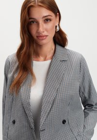 Kobieta z długimi rudymi włosami nosząca srebrne kolczyki, biały top i w kratkę marynarkę, z rękami w kieszeniach na jednolitym tle.