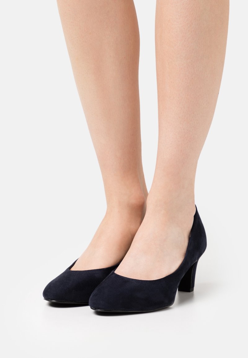 Jambes portant des chaussures en daim bleu marine à bout fermé et talon bloc bas sur fond blanc.