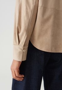 Beige longsleeve shirt met knoopmanchetten, gedragen met donkerblauwe denim broek, hand ontspannen aan de zijkant.