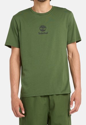 Homme portant un t-shirt Timberland vert olive et un short assorti, debout les bras le long du corps sur un fond uni.