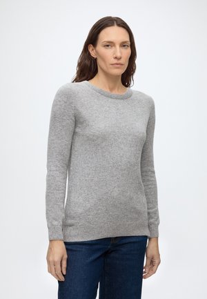 Femme aux longs cheveux bruns portant un pull gris clair à manches longues et un jean bleu foncé, debout devant un fond blanc uni.