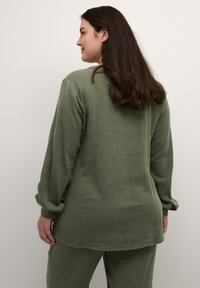 Olivgrünes, strukturiertes Stoffhemd mit langen Ärmeln, abgerundetem Saum und Knopfmanschetten. Das Design zeichnet sich durch eine lockere Passform und minimale Details aus.