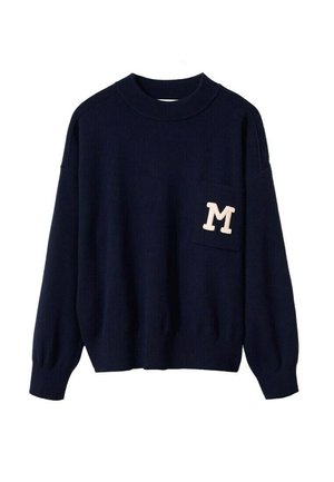 Femme portant un pull bleu marine avec des détails blancs, un pantalon en cuir noir taille haute et des bottines noires, debout avec une main dans la poche.