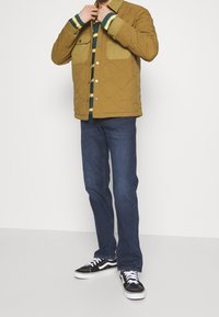 Gesteppte Senffarbene Jacke mit Fronttaschen, darüber ein grün-gelb gestreiftes Hemd, kombiniert mit dunkelblauen Jeans und schwarzen Turnschuhen.