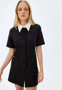 Vestido negro de manga corta con cuello blanco, frente de botones, dos bolsillos frontales y acentos decorativos de hardware. Textura de tela suave.