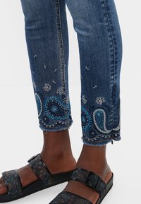 Jeans de denim bordados en azul oscuro, con diseños de paisley en azul y plata en el dobladillo, con bordes deshilachados y acentos plateados.