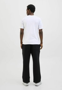 Weißes Baumwoll-T-Shirt mit kurzen Ärmeln, kombiniert mit schwarzen weit geschnittenen Hosen mit einer Gesäßtasche, vor einem neutralen Hintergrund.