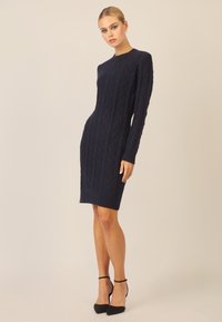 Marineblaues Strickkleid mit strukturiertem Diamantmuster, langen Ärmeln und rundem Ausschnitt, kombiniert mit schwarzen High Heels.