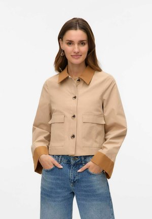 Giacca beige corta con colletto e polsini marroni, dotata di due tasche frontali e grandi bottoni, realizzata in tessuto liscio e leggero.