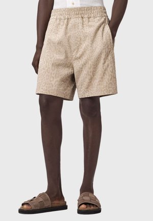 Mand iført beige leopardprint elastiske taljeshorts og brune vævede slidesandaler, står med hænderne i lommerne.