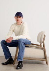 A light beige cable-knit sweater, blue denim jeans, and black shoes. Sitting on a wooden chair with a beige cushioned seat and backrest.