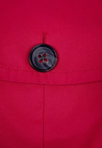 Röd tyg med en slät yta har en svart knapp i mitten, fyra hål med en sytråd i mitten och synlig sömdekoration.