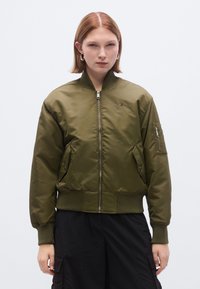 Χακί πράσινο bomber μπουφάν με φερμουάρ μπροστά, ριμπ μανσέτες, δύο πλαϊνές τσέπες και μικρό λογότυπο στο στήθος. Λείο, γυαλιστερό ύφασμα.