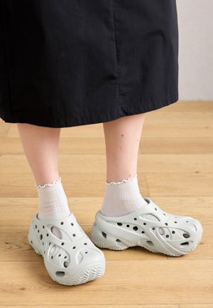 Füße in weißen Socken und hellgrauen Slip-on-Schuhen mit Lochmuster stehen auf einem Holzboden, darüber ist ein schwarzer Rock sichtbar.