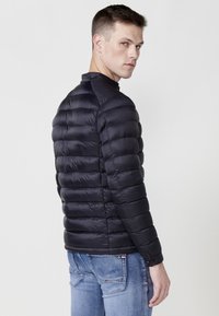 Chaqueta de plumas negra con secciones acolchadas horizontales, cuello alto y puños abotonados, combinada con jeans azul claro.