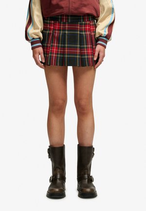 Personne portant une mini-jupe plissée à carreaux rouges, une veste bomber beige et bordeaux, et des bottes en cuir marron foncé mi-mollet avec des boucles.