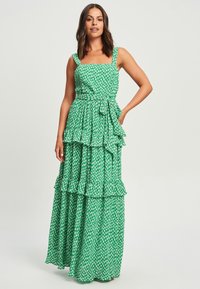 Zelena cvetna maksi obleka z nabranimi plastmi, kvadratnim izrezom in nastavljivim pasom na vezanje. Narejena iz lahko padajočega materiala z teksturiranim vzorcem.