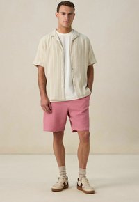 Korte mouwen, gestreepte crèmekleurige shirt boven een wit T-shirt, gecombineerd met gestructureerde roze shorts en witte sneakers met donkere accenten.