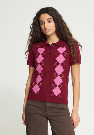 Jeune femme aux longs cheveux bouclés portant un pull bordeaux à manches courtes avec un motif losanges roses et un pantalon marron.