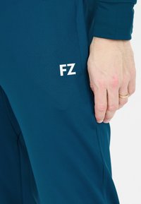 Teal idrottsbyxor med en slät textur, som har en vit "FZ"-logotyp på låret och en figurnära silhuett. Hand som rör vid tyget.