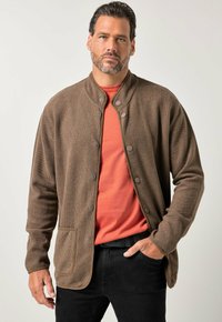 Brun strikket cardigan med rund krave, trykknapper og en forlomme, båret over en rød t-shirt, kombineret med sorte bukser.