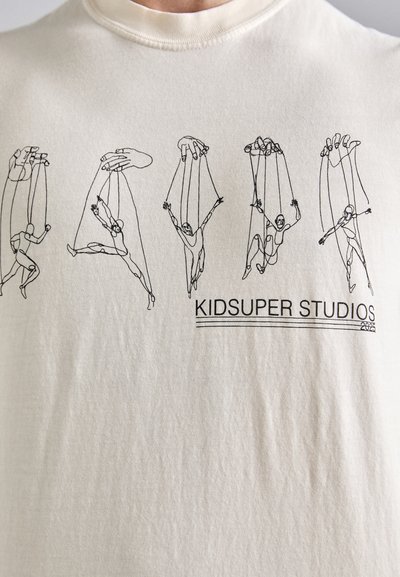 Cream-colored t-shirt featuring black line drawings of figures on strings, with "KIDSUPER STUDIOS 2025" printed below in black text.